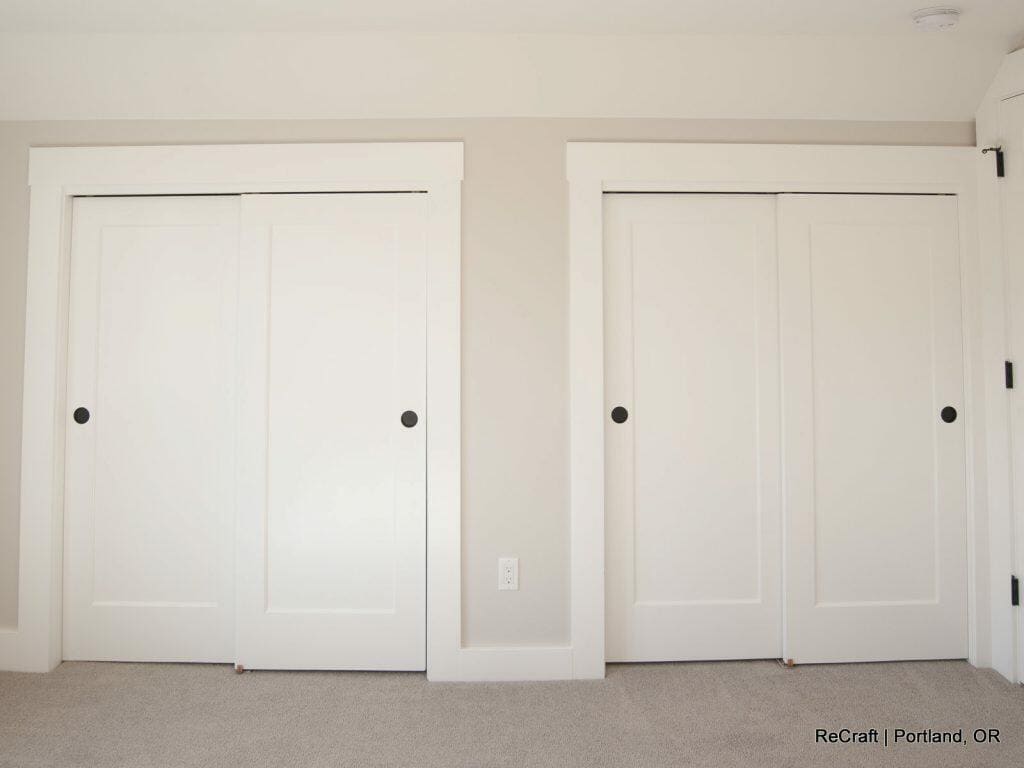 A room with two sets of white sliding closet doors, beige walls, and a carpeted floor. A small white power outlet is located between the closets. A room with two sets of white sliding closet doors, beige walls, and a carpeted floor. A small white power outlet is located between the closets.