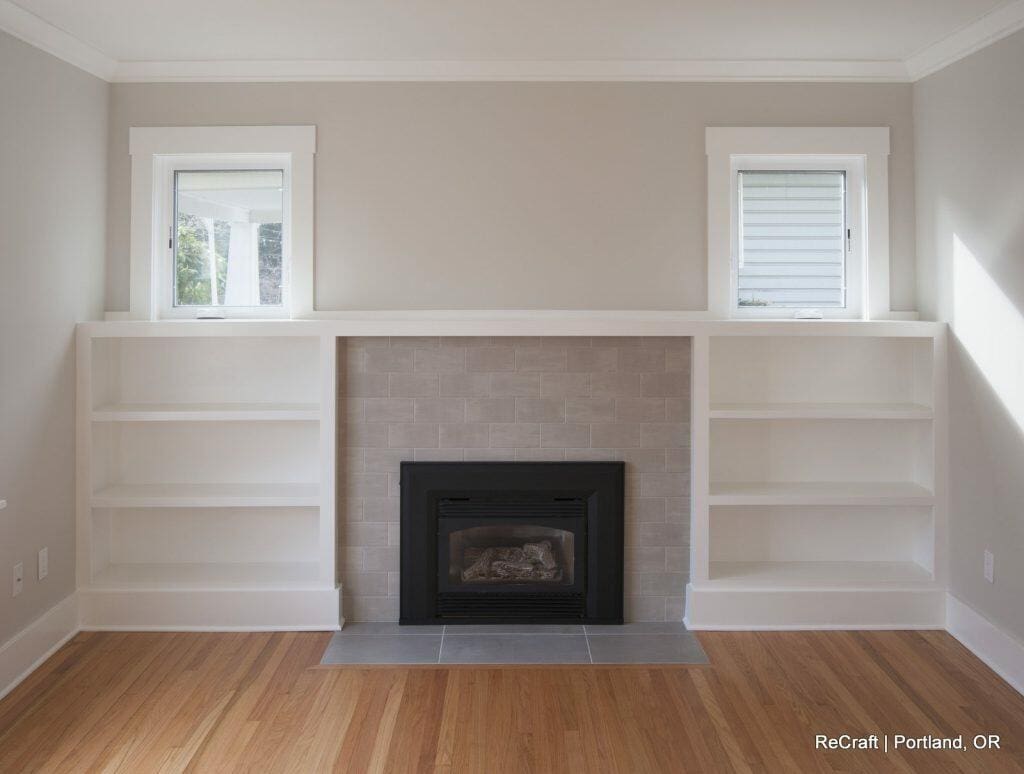 A living room features a fireplace with a tiled surround, flanked by two built-in bookcases. Two small windows are above the bookcases, and the floor is wooden. Text reads "ReCraft - Portland, OR. A living room features a fireplace with a tiled surround, flanked by two built-in bookcases. Two small windows are above the bookcases, and the floor is wooden. Text reads "ReCraft - Portland, OR.
