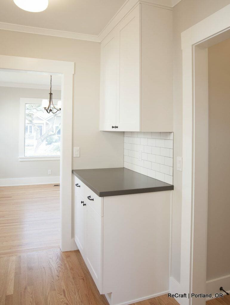 A modern kitchen corner with white cabinets, a dark countertop, and subway tile backsplash. Hardwood floor and a glimpse of a dining room with a window and chandelier are visible. A modern kitchen corner with white cabinets, a dark countertop, and subway tile backsplash. Hardwood floor and a glimpse of a dining room with a window and chandelier are visible.