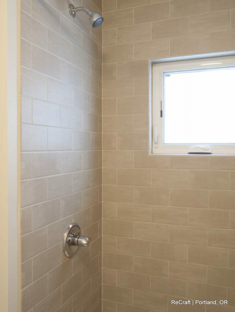 Shower area with beige subway tiles, a chrome showerhead, a control handle, and a small window on the right side allowing natural light. Shower area with beige subway tiles, a chrome showerhead, a control handle, and a small window on the right side allowing natural light.