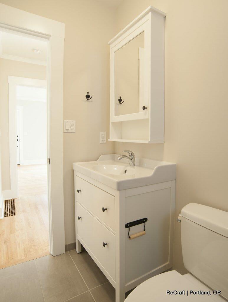 A small, clean bathroom with a white vanity, a built-in sink, a mirrored cabinet, and light grey tile flooring. A partially open door leads to a hallway with light wooden flooring. A small, clean bathroom with a white vanity, a built-in sink, a mirrored cabinet, and light grey tile flooring. A partially open door leads to a hallway with light wooden flooring.