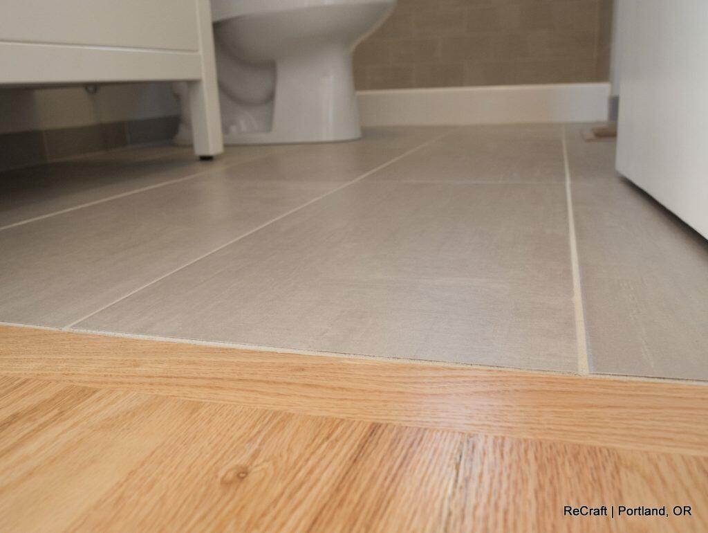 Close-up view showing the transition between a light wood floor and light gray tiled floor in a bathroom. A toilet is partially visible in the background. Close-up view showing the transition between a light wood floor and light gray tiled floor in a bathroom. A toilet is partially visible in the background.
