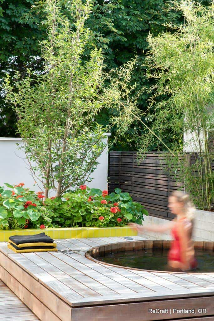 A small round hot tub is in a wooden deck surrounded by green plants and flowers. A person in a red outfit is moving near the tub. Text at the bottom reads, "ReCraft | Portland, OR. A small round hot tub is in a wooden deck surrounded by green plants and flowers. A person in a red outfit is moving near the tub. Text at the bottom reads, "ReCraft | Portland, OR.