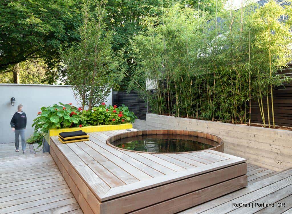 A modern outdoor space with a wooden deck, a circular soaking tub, a yellow bench with cushions, vibrant plants, and bamboo against the backdrop. A person is walking in the background. A modern outdoor space with a wooden deck, a circular soaking tub, a yellow bench with cushions, vibrant plants, and bamboo against the backdrop. A person is walking in the background.
