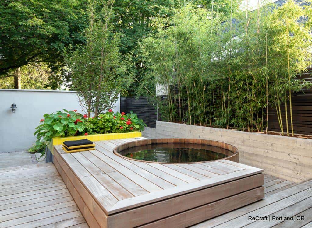 Wooden deck with circular hot tub, surrounded by greenery including bamboo and flowering plants, located in a backyard. Text on the image reads: "ReCraft | Portland, OR". Wooden deck with circular hot tub, surrounded by greenery including bamboo and flowering plants, located in a backyard. Text on the image reads: "ReCraft | Portland, OR".