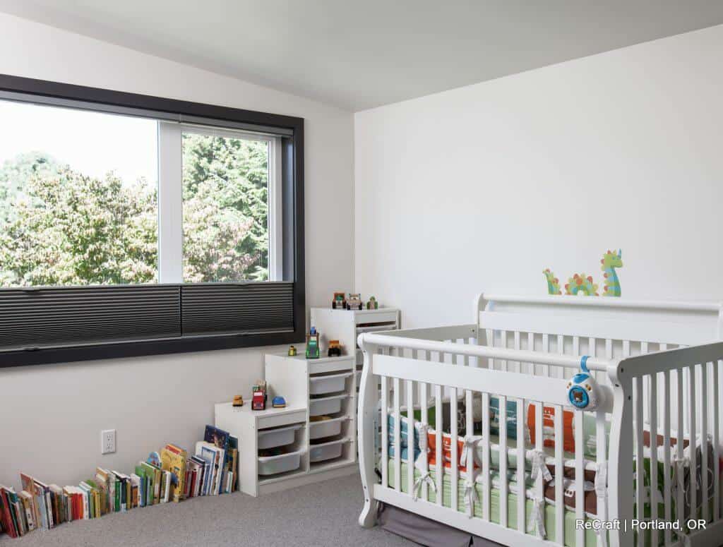 A nursery room with a white crib, shelves with bins and toys, a row of children's books, and a large window overlooking greenery. A nursery room with a white crib, shelves with bins and toys, a row of children's books, and a large window overlooking greenery.