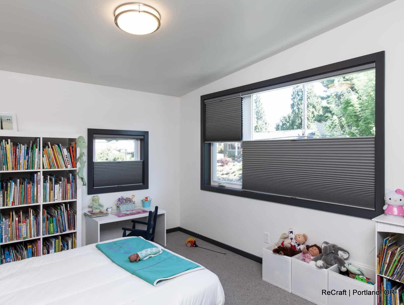 sorcinelli-3rd bed Brightly lit modern bedroom featuring large windows, a cozy bed with a turquoise blanket, a bookshelf filled with colorful books, and a white storage unit with toys, highlighting ReCraft Home Remodeling's design and renovation work in Portland.