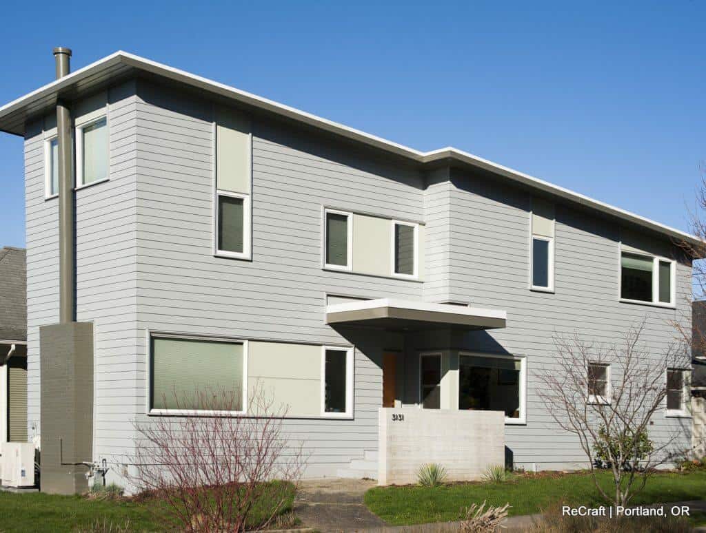 A two-story modern house with light gray horizontal siding, multiple rectangular windows, and a flat roof, set in a suburban neighborhood. Some shrubs and young trees are in the front yard. A two-story modern house with light gray horizontal siding, multiple rectangular windows, and a flat roof, set in a suburban neighborhood. Some shrubs and young trees are in the front yard.