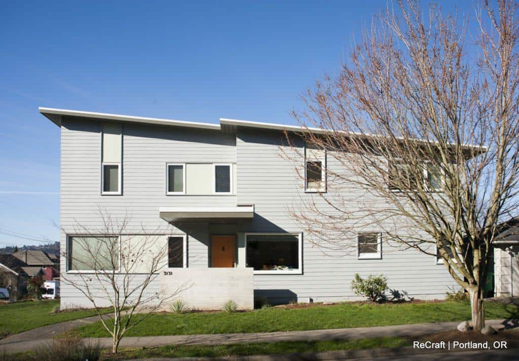 A two-story modern house with light grey siding, large windows, and a flat roof. A bare tree stands in the front yard. There's a small concrete porch with an orange door. The sky is clear. A two-story modern house with light grey siding, large windows, and a flat roof. A bare tree stands in the front yard. There's a small concrete porch with an orange door. The sky is clear.
