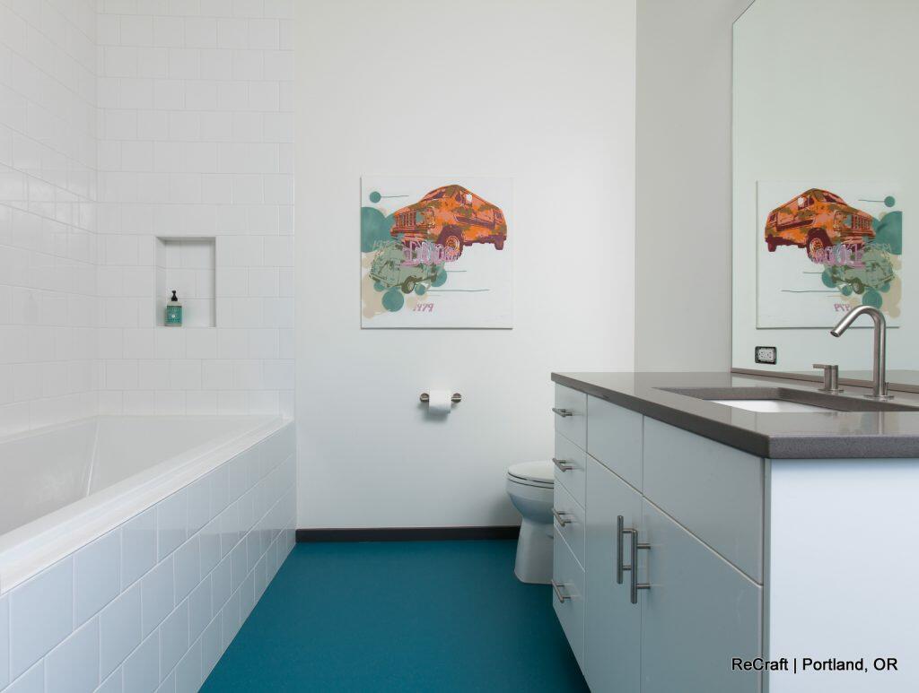 A modern bathroom with a bathtub on the left, a toilet in the center, a sink with a mirror on the right, and a painting on the wall. The floor is teal, and the walls are white. A modern bathroom with a bathtub on the left, a toilet in the center, a sink with a mirror on the right, and a painting on the wall. The floor is teal, and the walls are white.