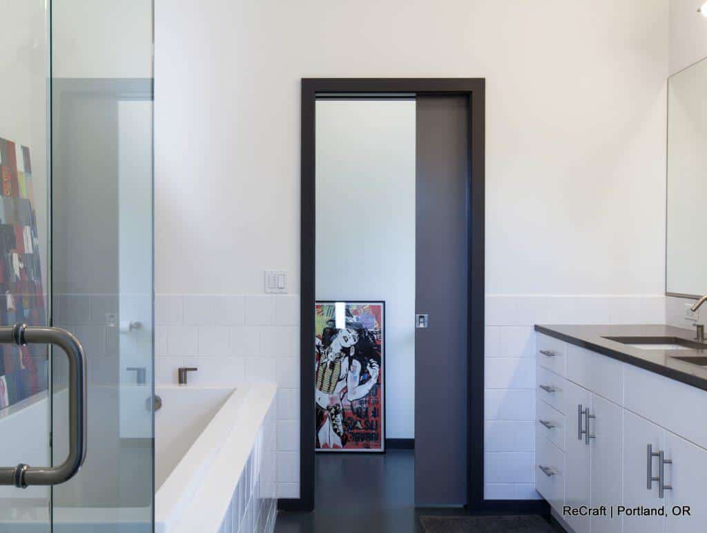 A modern bathroom featuring a bathtub on the left, a sliding door, and a countertop with a sink on the right. A framed pop art picture is partially visible behind the open door. A modern bathroom featuring a bathtub on the left, a sliding door, and a countertop with a sink on the right. A framed pop art picture is partially visible behind the open door.