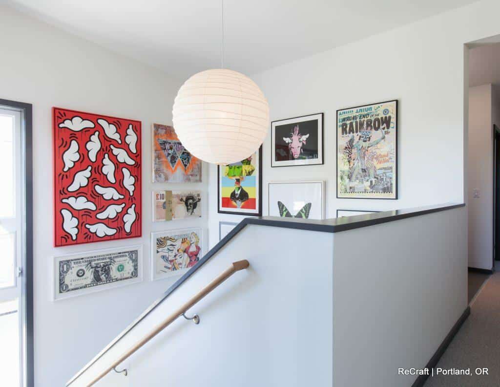 A staircase is adorned with various framed artworks, including a red piece with white shapes, a framed dollar bill, and several colorful prints. A spherical white light fixture hangs from the ceiling. A staircase is adorned with various framed artworks, including a red piece with white shapes, a framed dollar bill, and several colorful prints. A spherical white light fixture hangs from the ceiling.