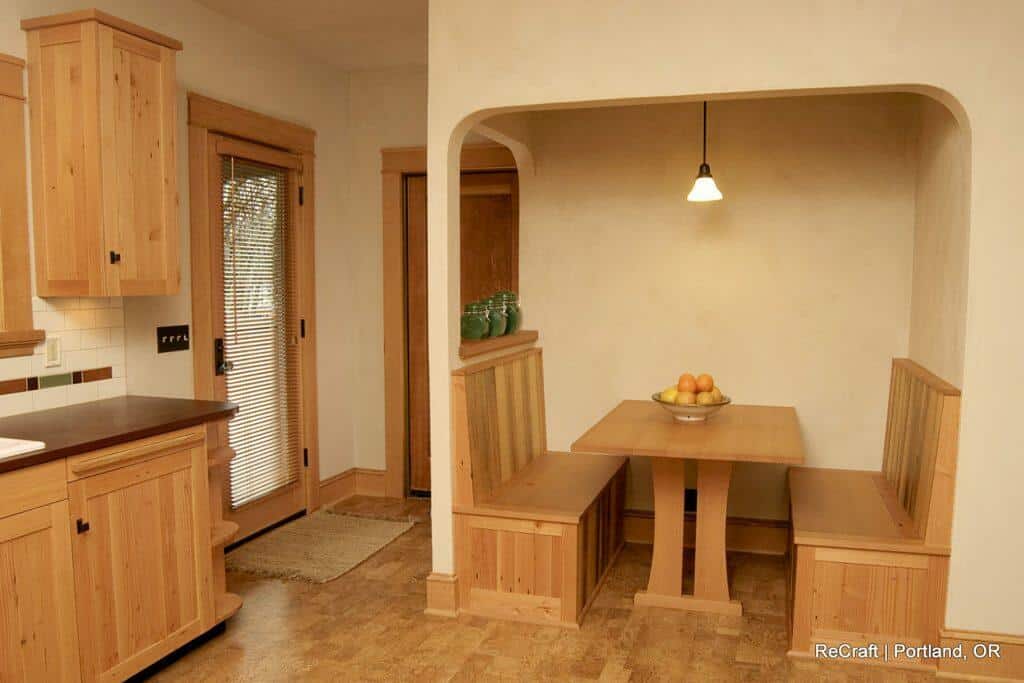 A small, wood-furnished kitchen nook with built-in benches, a table, a hanging light fixture, and a bowl of fruit. There are wooden cabinets and a door with blinds to the left. A small, wood-furnished kitchen nook with built-in benches, a table, a hanging light fixture, and a bowl of fruit. There are wooden cabinets and a door with blinds to the left.