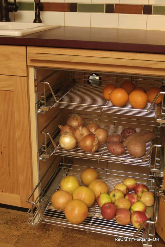 A kitchen cabinet with pull-out wire baskets containing onions, potatoes, sweet potatoes, oranges, apples, and other fruits and vegetables beside a sink with a dark countertop. A kitchen cabinet with pull-out wire baskets containing onions, potatoes, sweet potatoes, oranges, apples, and other fruits and vegetables beside a sink with a dark countertop.