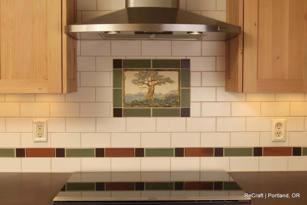 A kitchen backsplash with white subway tiles, featuring a central decorative tile with a tree, under a stainless steel range hood. Light wood cabinets are on either side. A kitchen backsplash with white subway tiles, featuring a central decorative tile with a tree, under a stainless steel range hood. Light wood cabinets are on either side.