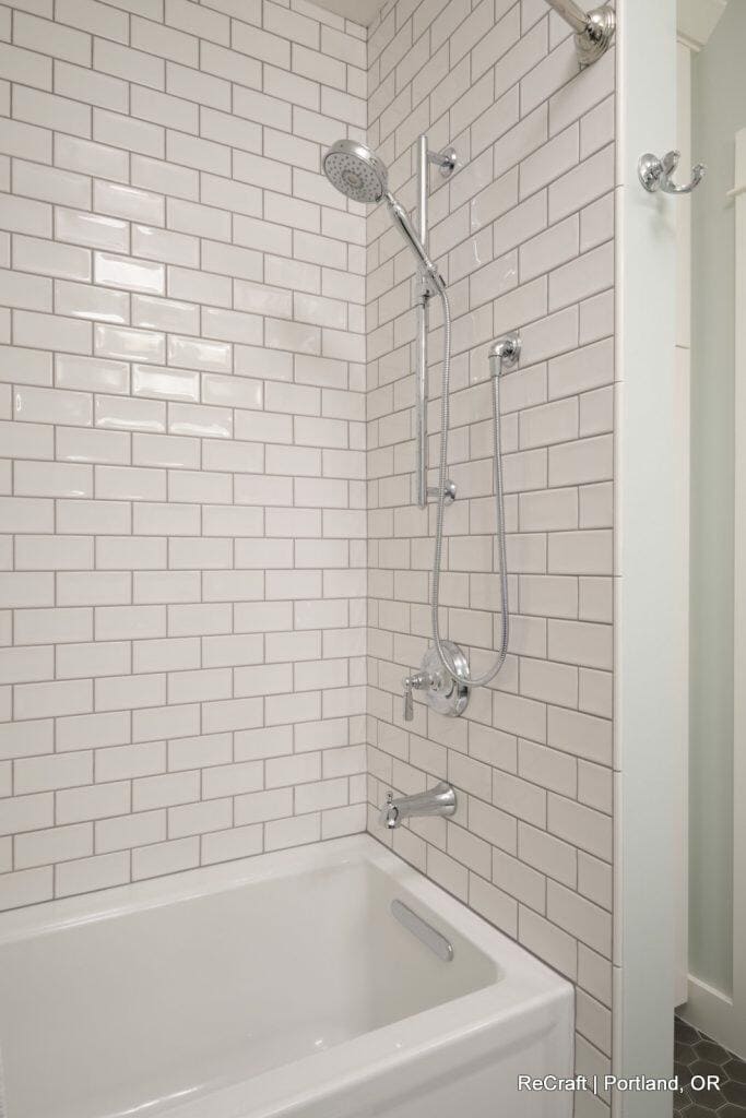 A white-tiled bathroom features a showerhead, handheld shower, and bathtub with a built-in soap holder. The tiles are arranged in a classic subway pattern. A hook and towel can be seen on the far right. A white-tiled bathroom features a showerhead, handheld shower, and bathtub with a built-in soap holder. The tiles are arranged in a classic subway pattern. A hook and towel can be seen on the far right.