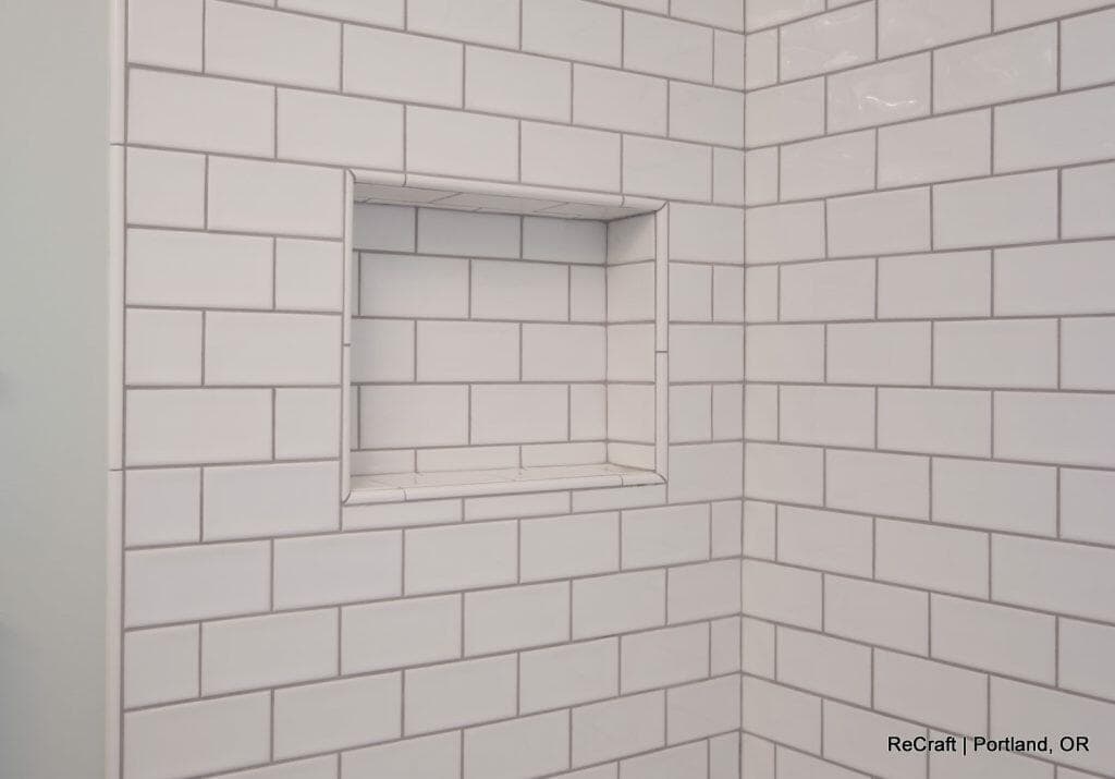 Close-up of a tiled shower niche with white subway tiles and light gray grout lines, framed by matching tiles and located in a corner. Close-up of a tiled shower niche with white subway tiles and light gray grout lines, framed by matching tiles and located in a corner.