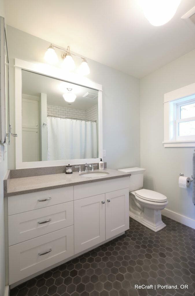 A bright bathroom with a large mirror above a white vanity, a gray countertop, a toilet, a towel on a rack, and a partially visible shower with a white curtain. The floor has hexagonal dark gray tiles. A bright bathroom with a large mirror above a white vanity, a gray countertop, a toilet, a towel on a rack, and a partially visible shower with a white curtain. The floor has hexagonal dark gray tiles.