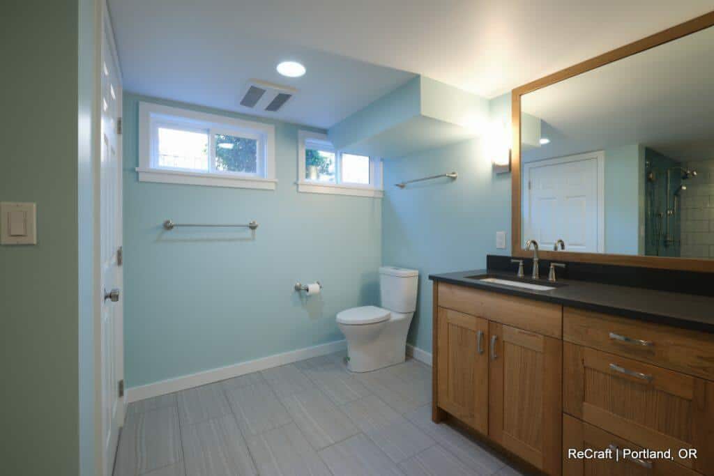 A spacious, modern bathroom with light blue walls, a large mirror above a wooden vanity, a toilet, and a walk-in shower. Two small windows provide natural light. Text in lower right: "ReCraft | Portland, OR". A spacious, modern bathroom with light blue walls, a large mirror above a wooden vanity, a toilet, and a walk-in shower. Two small windows provide natural light. Text in lower right: "ReCraft | Portland, OR".