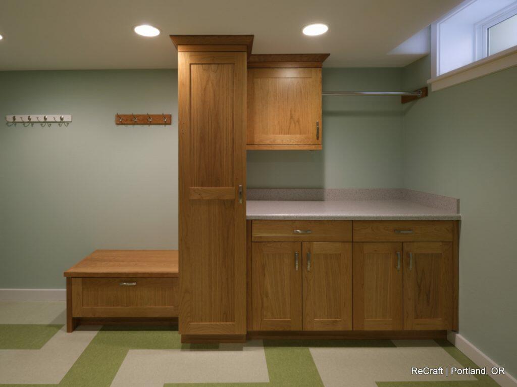 A room with light green walls, a wooden cabinet and shelving unit, a bench with hooks above it, and a countertop with storage below. The floor has a green and white checkered pattern. A room with light green walls, a wooden cabinet and shelving unit, a bench with hooks above it, and a countertop with storage below. The floor has a green and white checkered pattern.