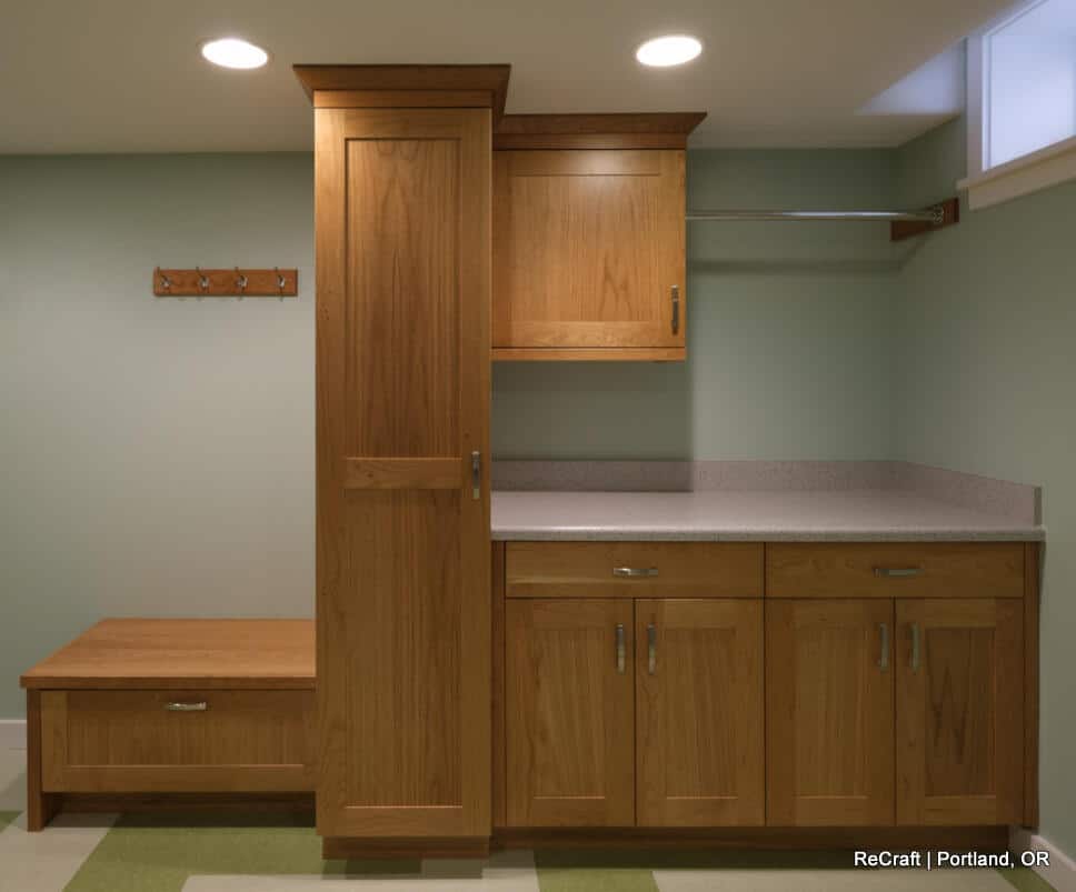 A wooden built-in storage unit with multiple cabinets and drawers is set against a light green wall. It includes a bench with a drawer below and a hook rack above. The flooring is patterned. A wooden built-in storage unit with multiple cabinets and drawers is set against a light green wall. It includes a bench with a drawer below and a hook rack above. The flooring is patterned.