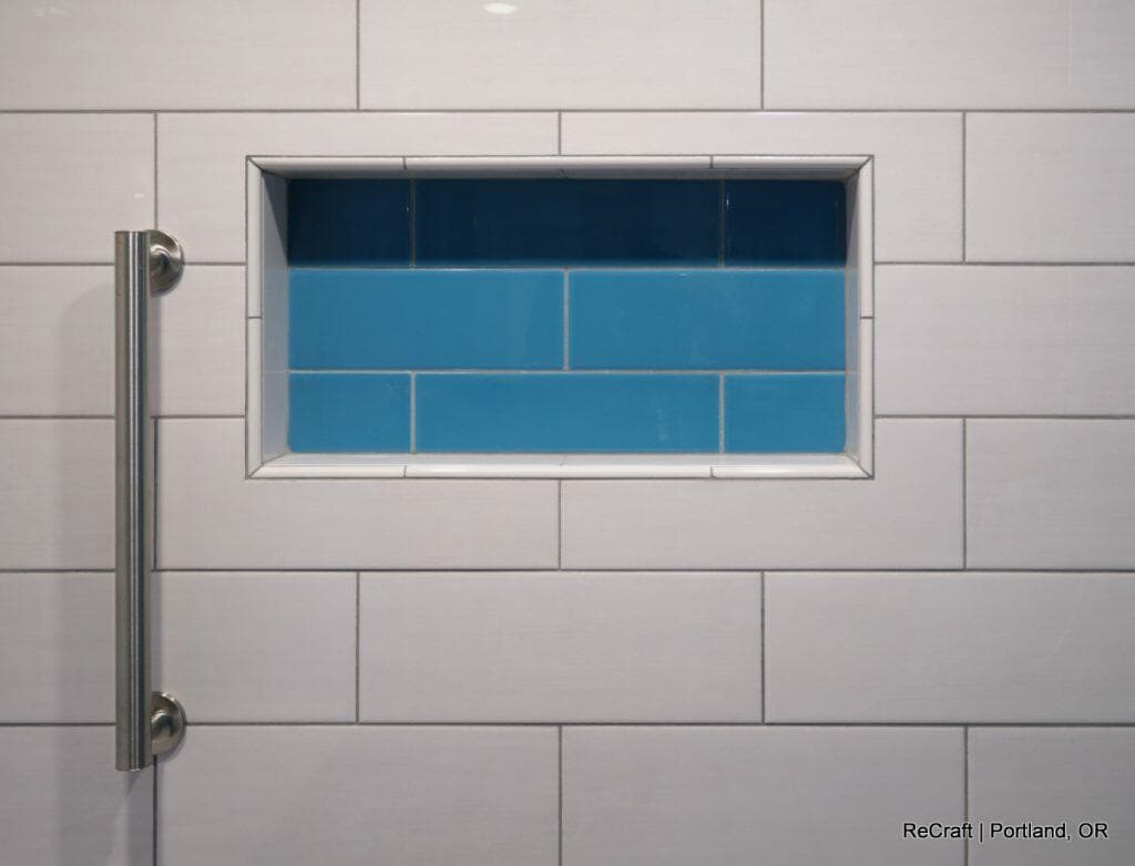 A wall of white rectangular tiles features a recessed niche with blue tiles in a shower. A stainless steel handle is mounted vertically on the left side of the wall. A wall of white rectangular tiles features a recessed niche with blue tiles in a shower. A stainless steel handle is mounted vertically on the left side of the wall.