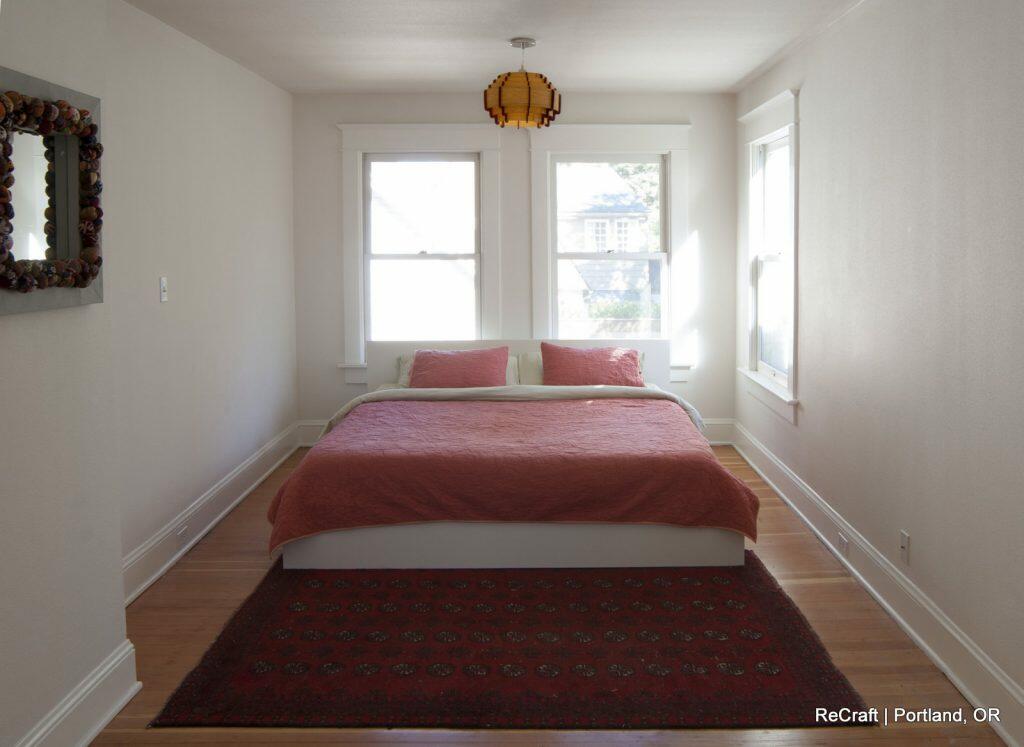 A minimalist bedroom with a white bed frame, red bedding, two windows, a decorative mirror on the left wall, a pendant light, and a patterned red rug on the wooden floor. Text: "ReCraft | Portland, OR". A minimalist bedroom with a white bed frame, red bedding, two windows, a decorative mirror on the left wall, a pendant light, and a patterned red rug on the wooden floor. Text: "ReCraft | Portland, OR".