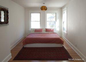 A minimalist bedroom with a white bed frame, red bedding, two windows, a decorative mirror on the left wall, a pendant light, and a patterned red rug on the wooden floor. Text: 