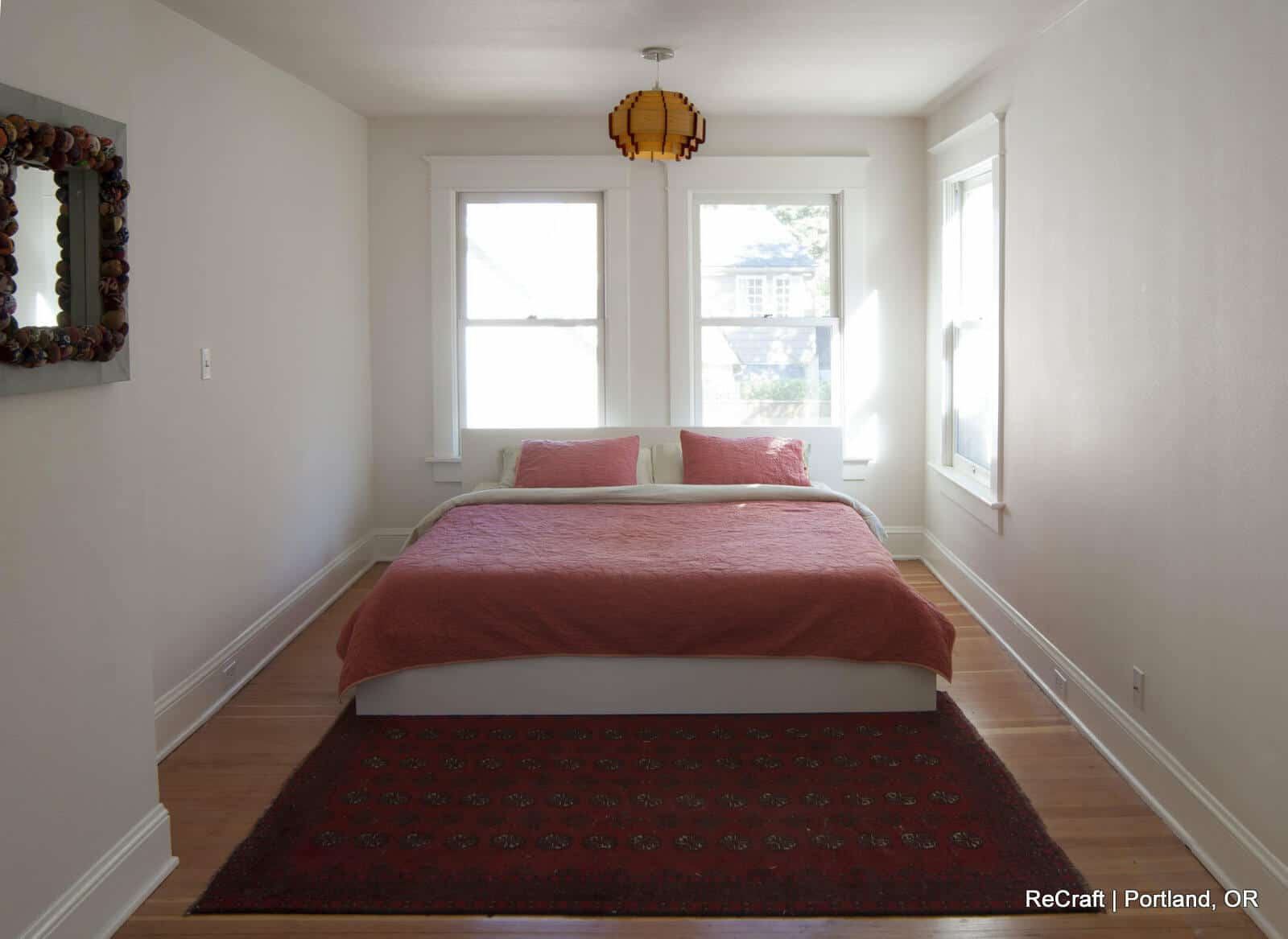 A minimalist bedroom with a white bed frame, red bedding, two windows, a decorative mirror on the left wall, a pendant light, and a patterned red rug on the wooden floor. Text: