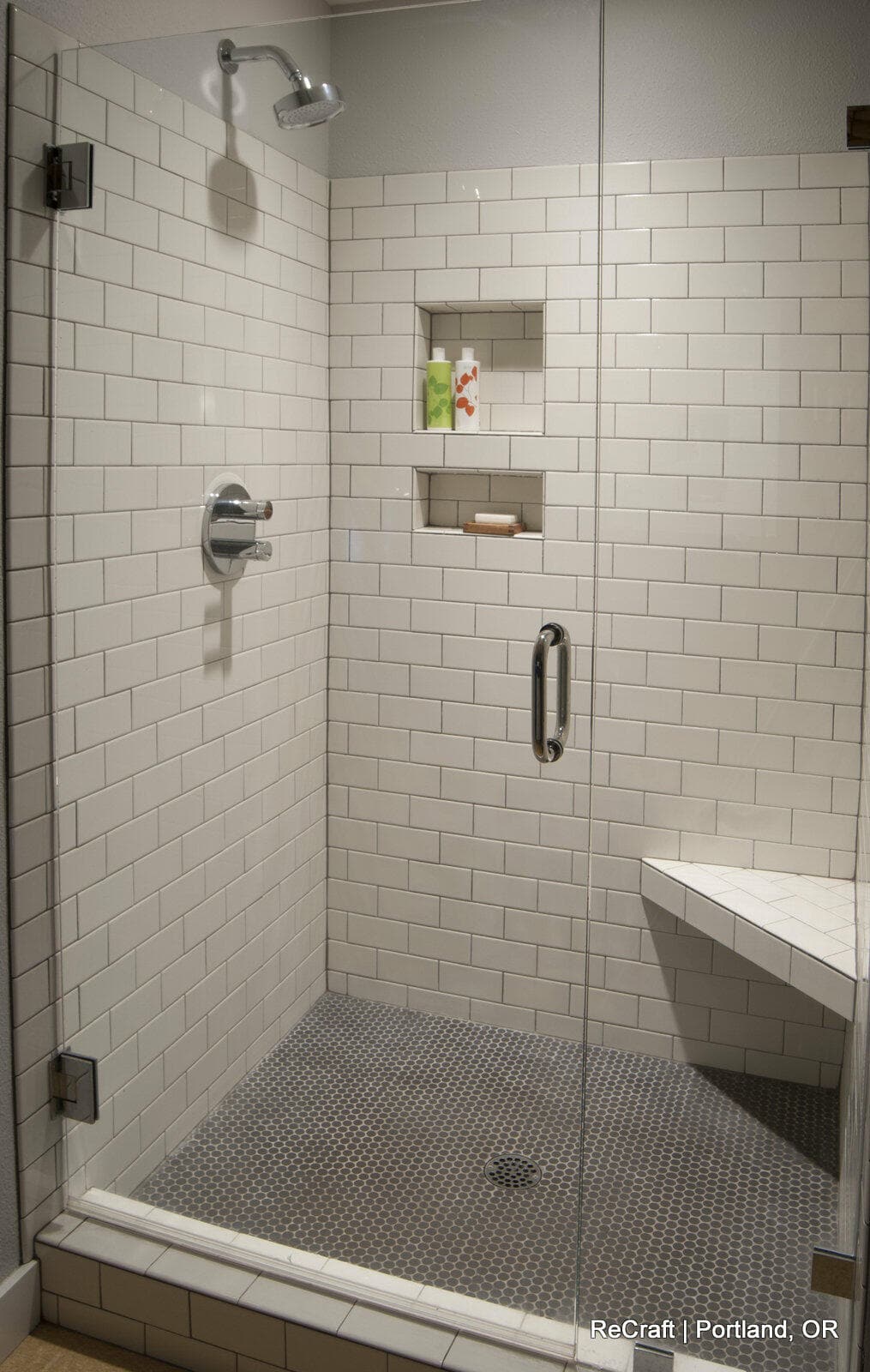 A modern shower with white subway tiles, a glass door, two recessed shelves with toiletries, a chrome showerhead, and a built-in bench.