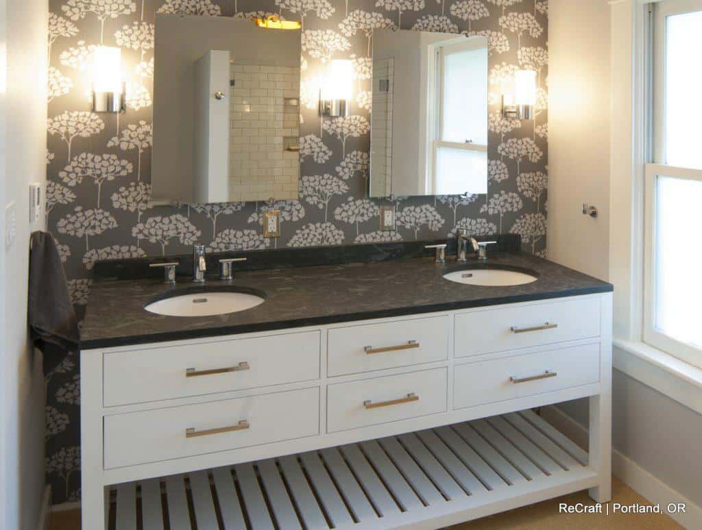 Modern bathroom with double sinks, black countertop, mirrored cabinets, and wall-mounted lights. White vanity has six drawers and an open shelf below. Grey floral-patterned wallpaper on accent wall. Modern bathroom with double sinks, black countertop, mirrored cabinets, and wall-mounted lights. White vanity has six drawers and an open shelf below. Grey floral-patterned wallpaper on accent wall.