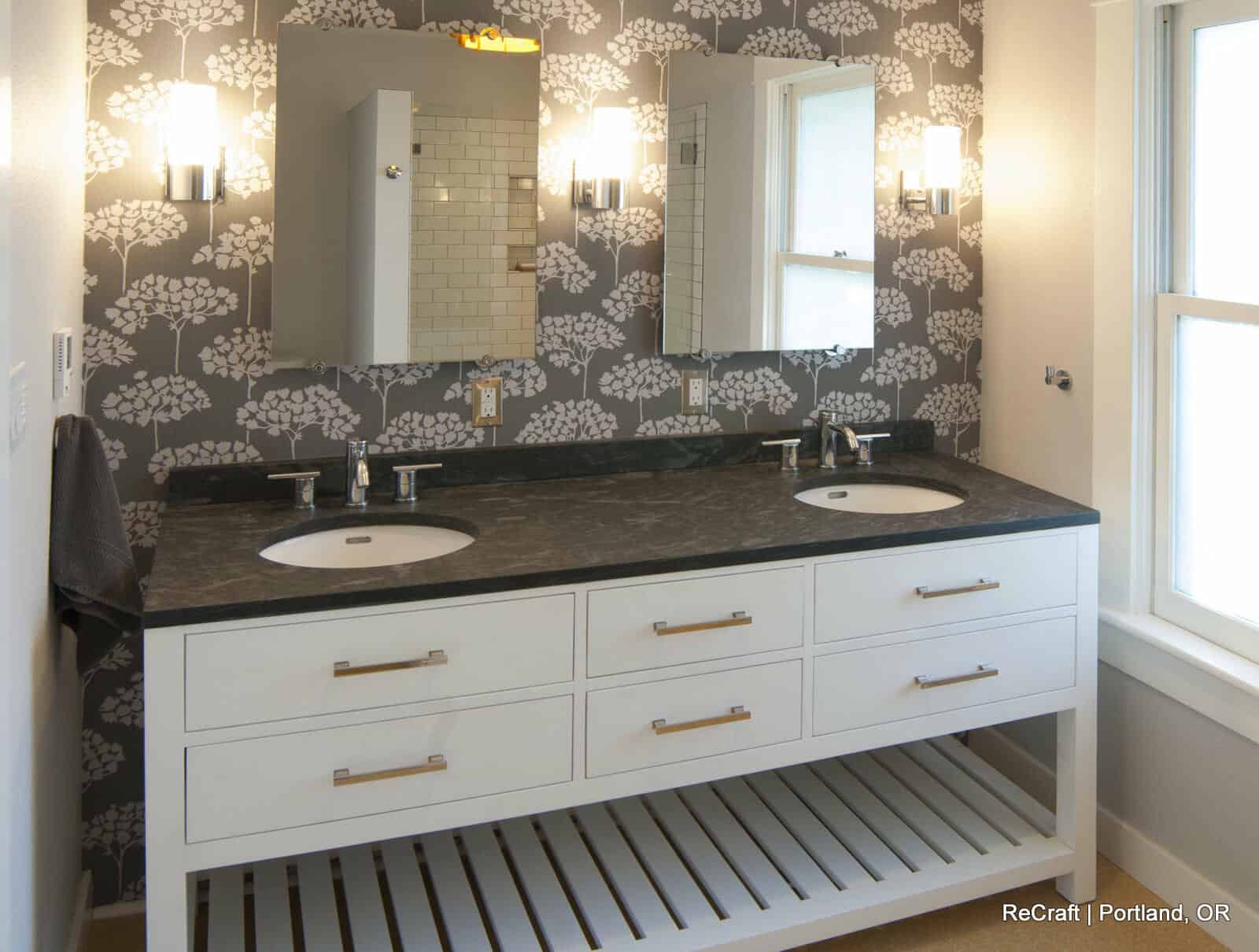 Modern bathroom with double sinks, black countertop, mirrored cabinets, and wall-mounted lights. White vanity has six drawers and an open shelf below. Grey floral-patterned wallpaper on accent wall.