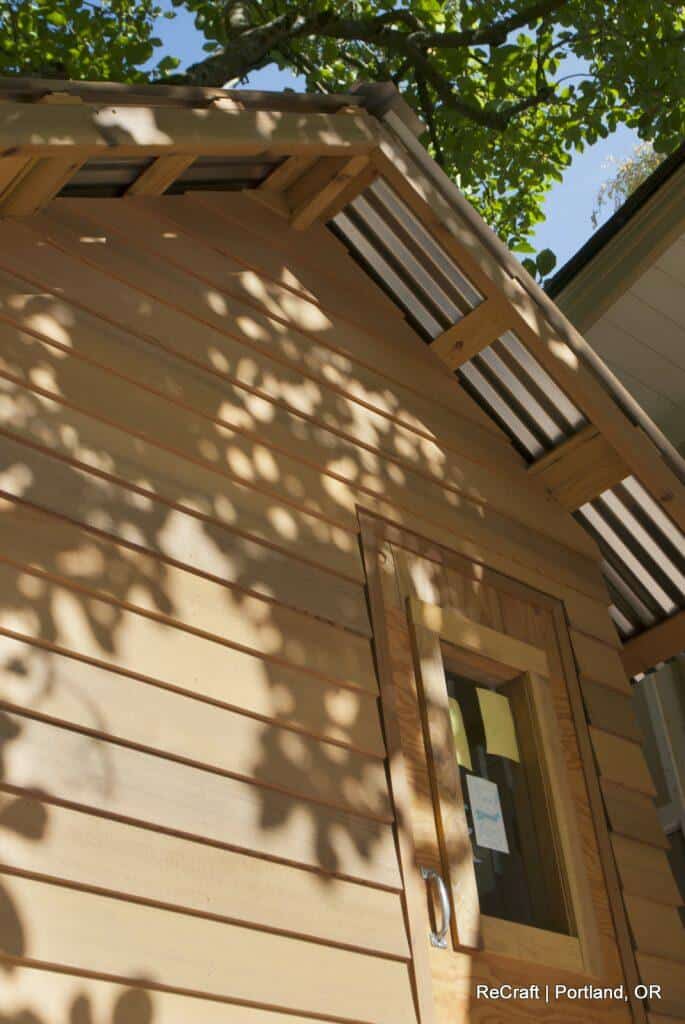 Close-up view of the side of a wooden structure with a door and translucent panels on the roof. Sunlight filters through tree leaves, casting shadows on the wall. Text at the bottom right: "ReCraft | Portland, OR. Close-up view of the side of a wooden structure with a door and translucent panels on the roof. Sunlight filters through tree leaves, casting shadows on the wall. Text at the bottom right: "ReCraft | Portland, OR.