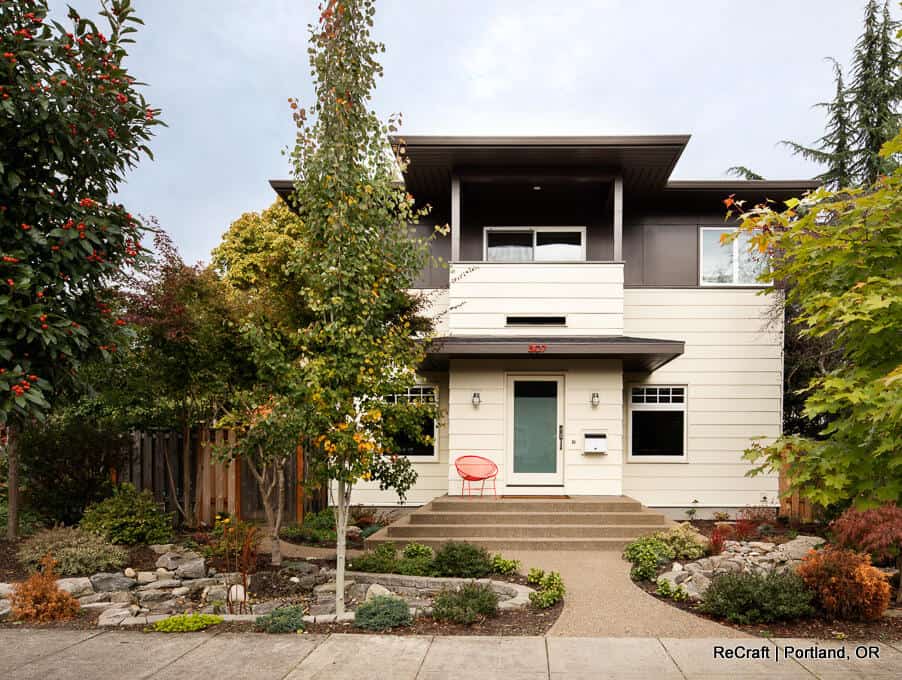 Two-story modern house with beige and dark gray exterior, surrounded by landscaped greenery. Red chair on front porch, steps leading to the entrance. Located in a suburban neighborhood. Two-story modern house with beige and dark gray exterior, surrounded by landscaped greenery. Red chair on front porch, steps leading to the entrance. Located in a suburban neighborhood.