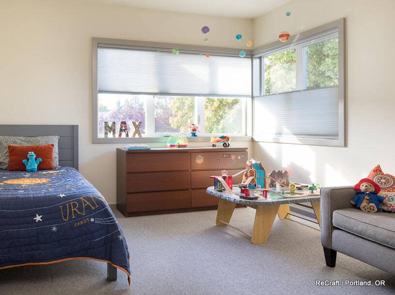 A child's bedroom with a bed, dresser, and toys. The bed has a blue space-themed blanket. A window lets in natural light, and a toy table is set up near a gray chair. A child's bedroom with a bed, dresser, and toys. The bed has a blue space-themed blanket. A window lets in natural light, and a toy table is set up near a gray chair.