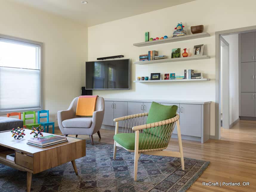 A modern living room with a TV on the wall, bookshelves, an armchair, a green chair, a wooden coffee table, and a patterned rug on a wooden floor. Kids' chairs are near a window. A modern living room with a TV on the wall, bookshelves, an armchair, a green chair, a wooden coffee table, and a patterned rug on a wooden floor. Kids' chairs are near a window.