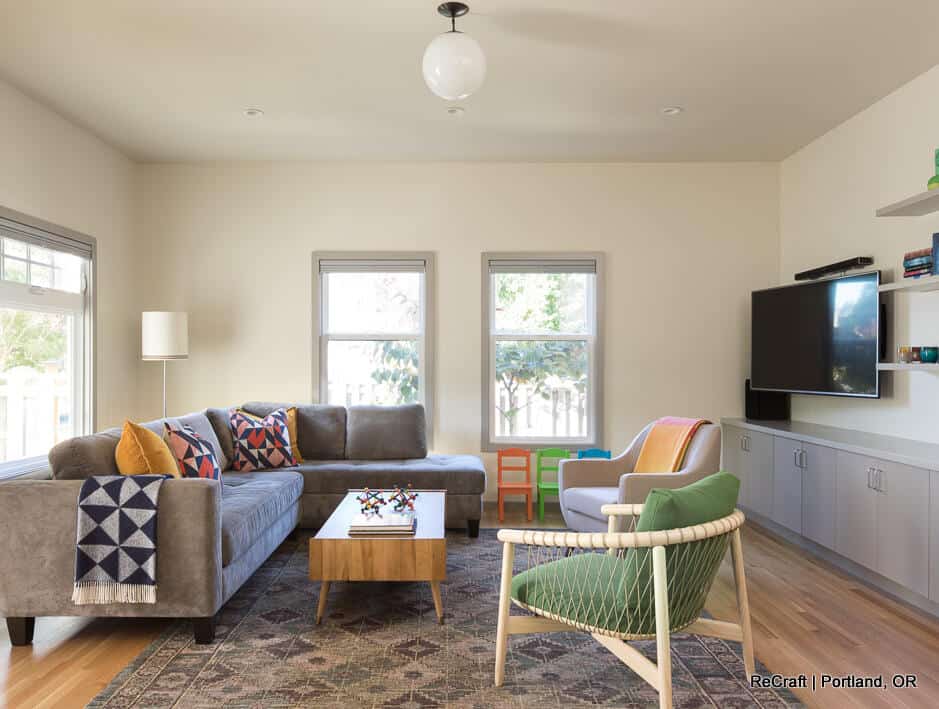 A modern living room with a large gray sofa, a green armchair, a wooden coffee table, and a wall-mounted TV. The room has two windows, a light fixture, and light-colored walls. A modern living room with a large gray sofa, a green armchair, a wooden coffee table, and a wall-mounted TV. The room has two windows, a light fixture, and light-colored walls.