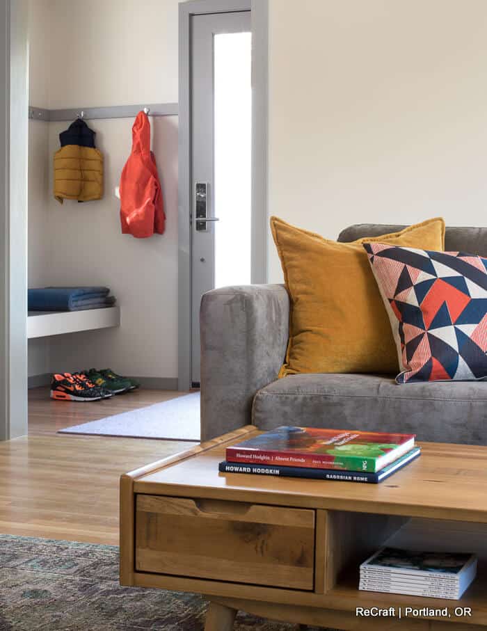 A cozy living room with a gray couch, colorful pillows, and a wooden coffee table. A book and magazine rest on the table. In the background, coats hang by a door with shoes lined up below. A cozy living room with a gray couch, colorful pillows, and a wooden coffee table. A book and magazine rest on the table. In the background, coats hang by a door with shoes lined up below.