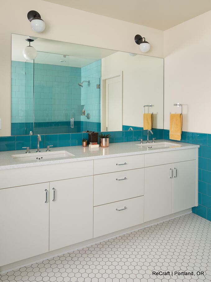 A modern bathroom with white cabinets, two sinks, a large mirror, turquoise wall tiles, and white hexagon floor tiles. Two yellow hand towels and copper accessories are on the counter. A modern bathroom with white cabinets, two sinks, a large mirror, turquoise wall tiles, and white hexagon floor tiles. Two yellow hand towels and copper accessories are on the counter.