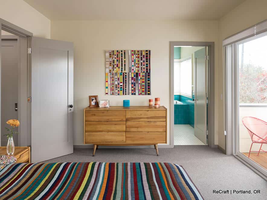 A bedroom with a striped bedspread, wooden dresser, modern artwork, two doors, and a glass sliding door leading to a balcony. The dresser has various items on top. A bedroom with a striped bedspread, wooden dresser, modern artwork, two doors, and a glass sliding door leading to a balcony. The dresser has various items on top.