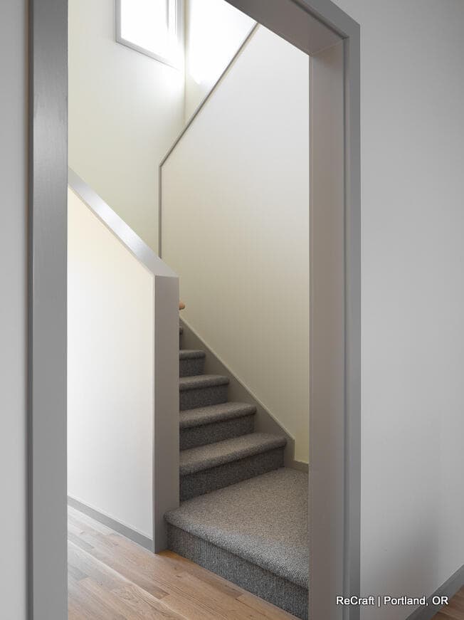 A carpeted staircase with gray handrails leading upwards, viewed through a doorway. The space is lit by natural light from a window at the top. Flooring transitions from wood to carpet. A carpeted staircase with gray handrails leading upwards, viewed through a doorway. The space is lit by natural light from a window at the top. Flooring transitions from wood to carpet.