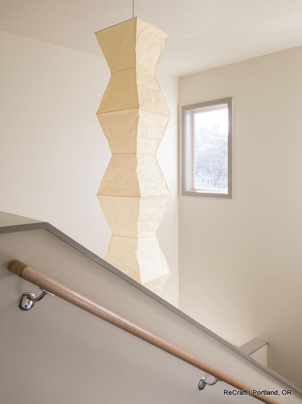A staircase with a wooden handrail is next to a window and a large, beige, geometric hanging light fixture. Text at the bottom right reads "ReCraft | Portland, OR. A staircase with a wooden handrail is next to a window and a large, beige, geometric hanging light fixture. Text at the bottom right reads "ReCraft | Portland, OR.