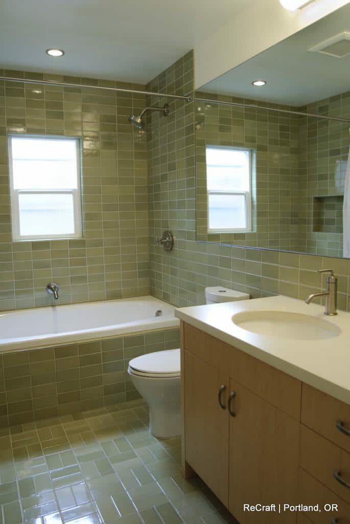 Modern bathroom with green tile walls and flooring, a bathtub, a toilet, and a sink. A large mirror is above the sink, and two frosted windows allow light into the room. Modern bathroom with green tile walls and flooring, a bathtub, a toilet, and a sink. A large mirror is above the sink, and two frosted windows allow light into the room.