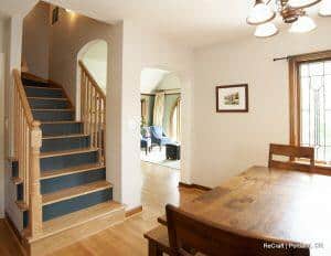 A brightly lit dining area with a wooden table and chairs, adjacent to a staircase with wooden railings and arched doorways leading to a living room.