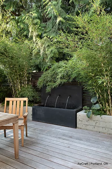 Modern outdoor space featuring a sleek black water feature, surrounded by lush greenery and bamboo, with a wooden chair on a deck, showcasing ReCraft Home Remodeling's design expertise in Portland.