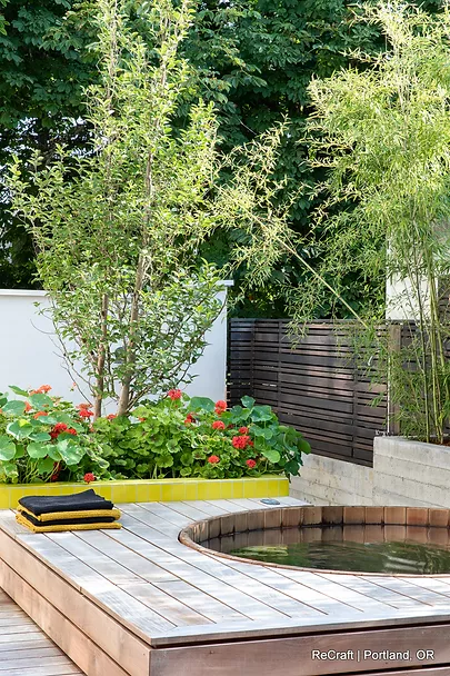 Modern outdoor garden space featuring a circular pond, wooden deck, vibrant flowers, and lush greenery, showcasing ReCraft Home Remodeling's landscaping and design capabilities.