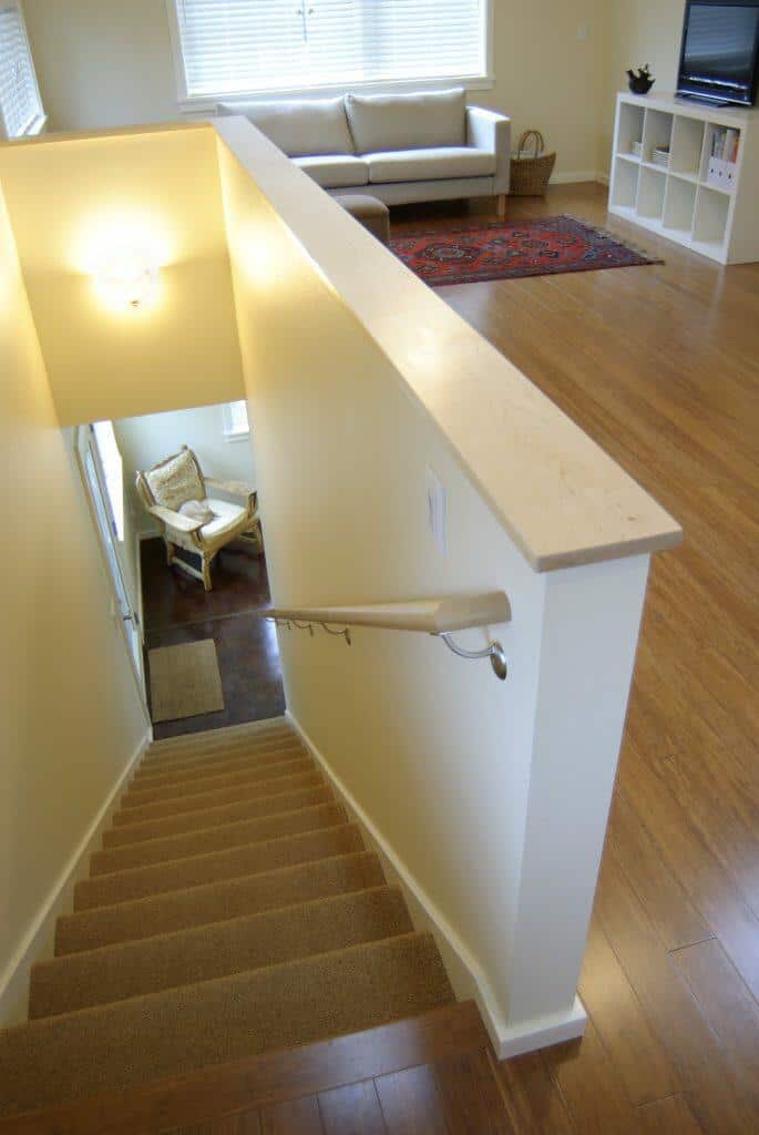 Staircase with a wooden handrail leading to an upper floor with a beige sofa, TV, red rug, and bookshelf. Lower area has a small seating nook with a chair near a window. Staircase with a wooden handrail leading to an upper floor with a beige sofa, TV, red rug, and bookshelf. Lower area has a small seating nook with a chair near a window.