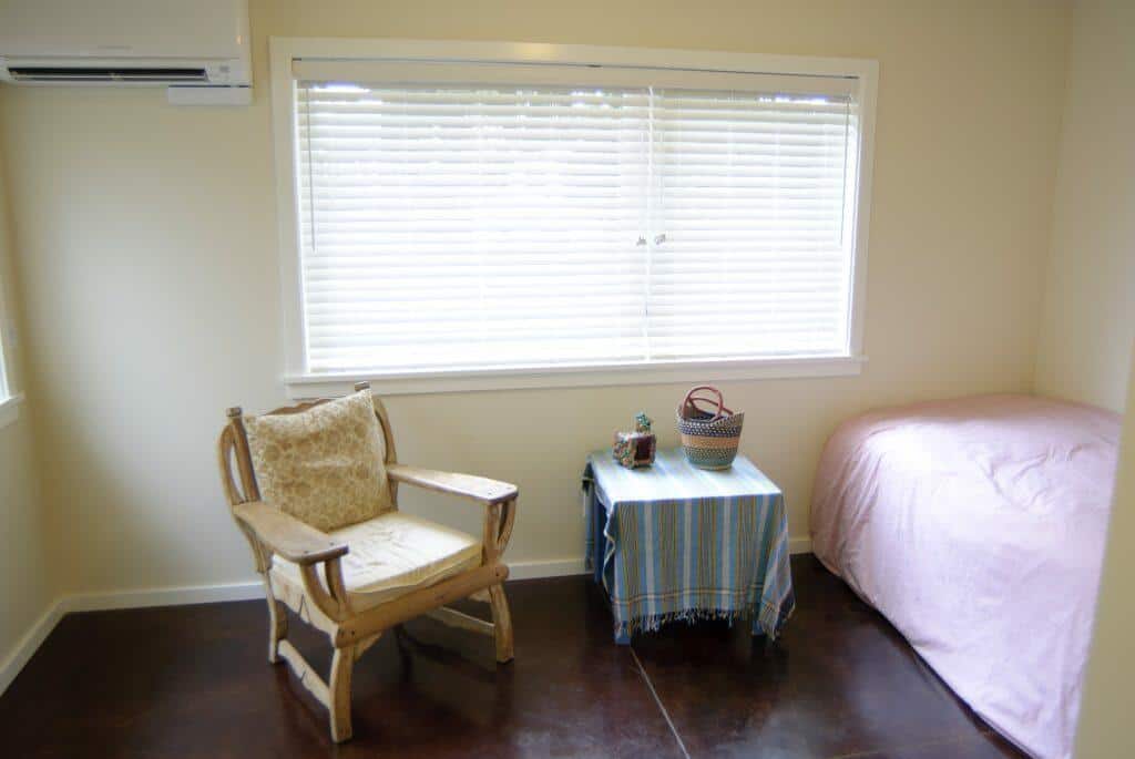 A small room with a chair, a bed with a pink blanket, and a small table covered with a blue cloth. There is a window with closed blinds and an air conditioner unit above it. A small room with a chair, a bed with a pink blanket, and a small table covered with a blue cloth. There is a window with closed blinds and an air conditioner unit above it.