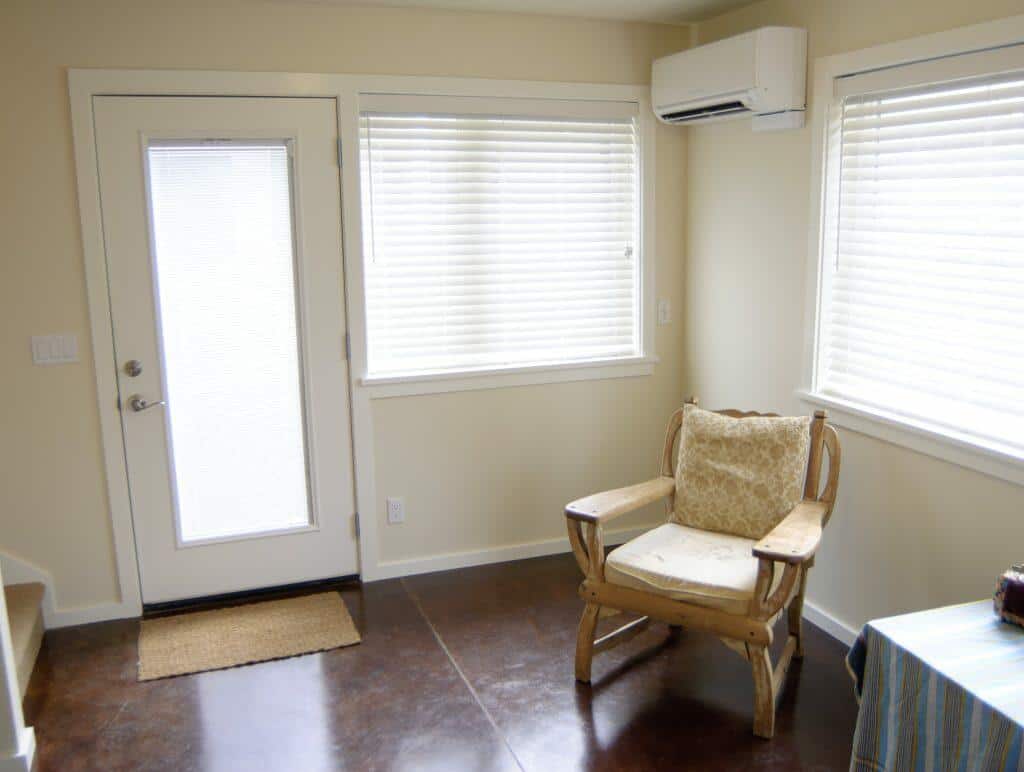 A small room featuring a windowed door, a window with white blinds, a wooden chair with a cushion, and an air conditioning unit above the window. A small room featuring a windowed door, a window with white blinds, a wooden chair with a cushion, and an air conditioning unit above the window.