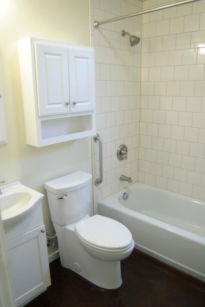 A bathroom features a white toilet, bathtub with an overhead shower, a sink with a white cabinet, and a wall-mounted white storage unit. The tile background is white. A bathroom features a white toilet, bathtub with an overhead shower, a sink with a white cabinet, and a wall-mounted white storage unit. The tile background is white.